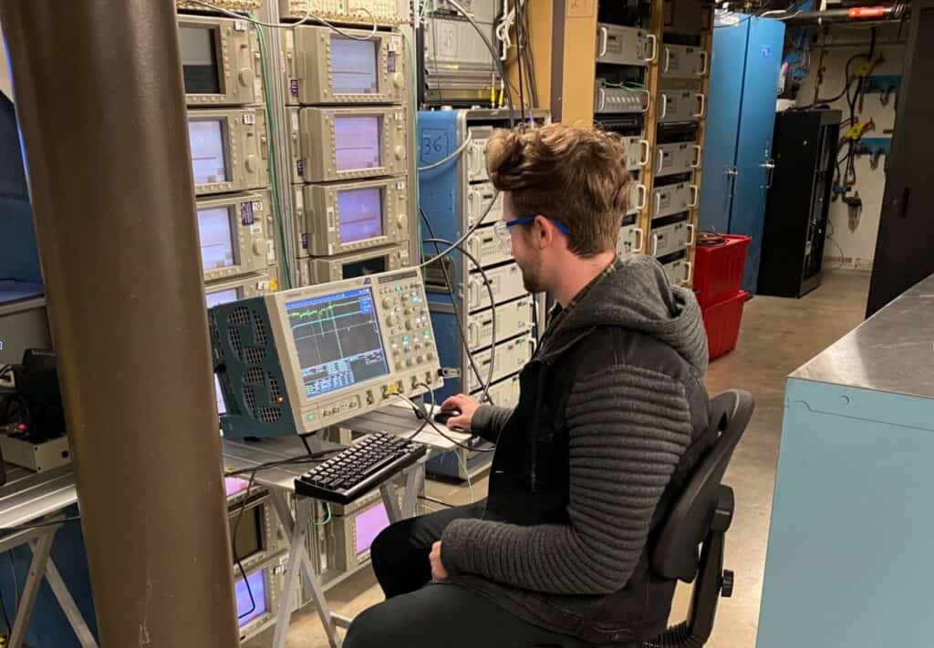 Landon, a student researcher, sits at a workstation in the Plasma and Pulsed Power Laboratory reviewing signals on an oscilloscope. He wears safety glasses and is surrounded by racks of diagnostic instruments and stacked monitors used for pulsed power experiments.